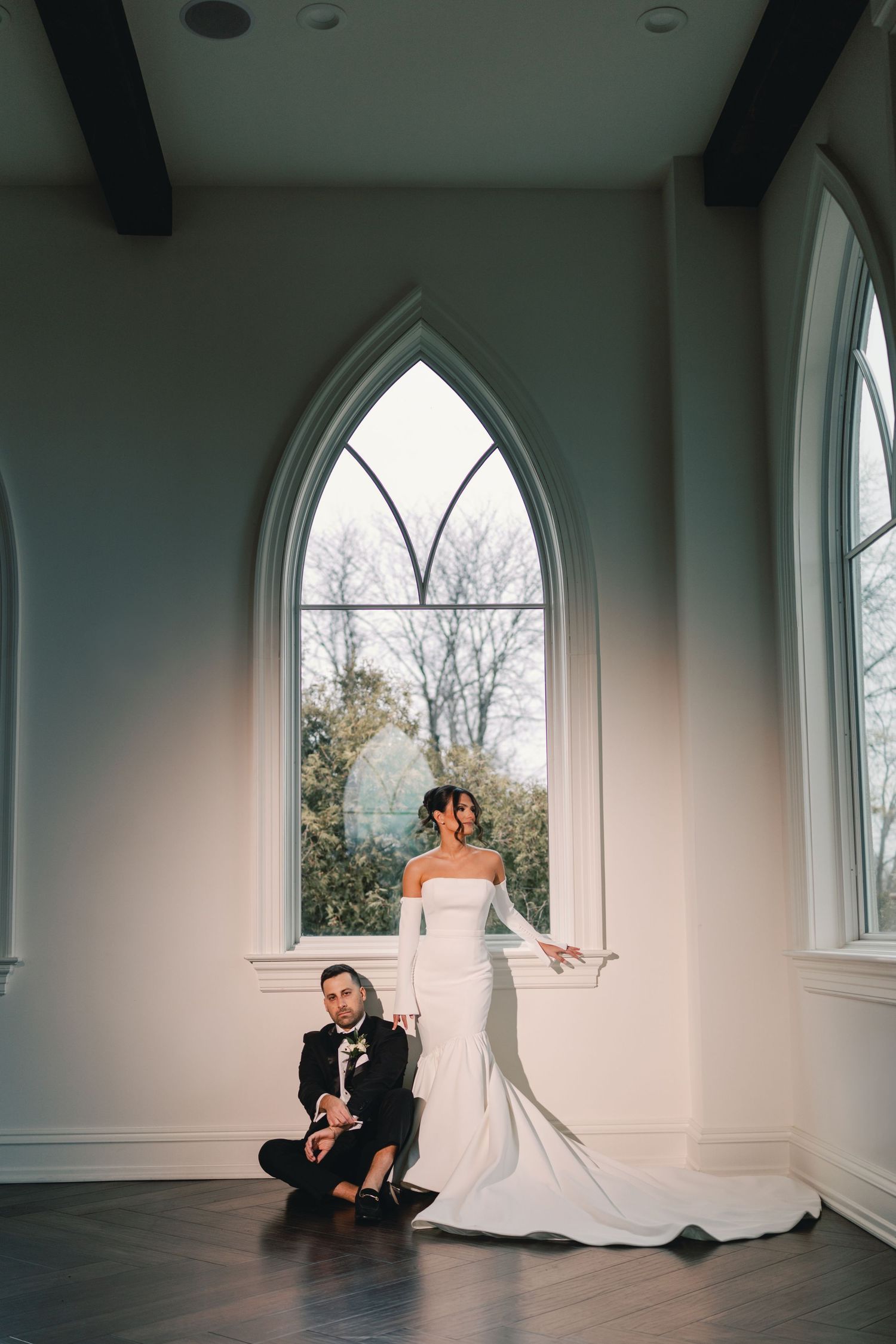 Bride in off-shoulder gown and groom sitting before tall Gothic arched windows in a stunning chapel.