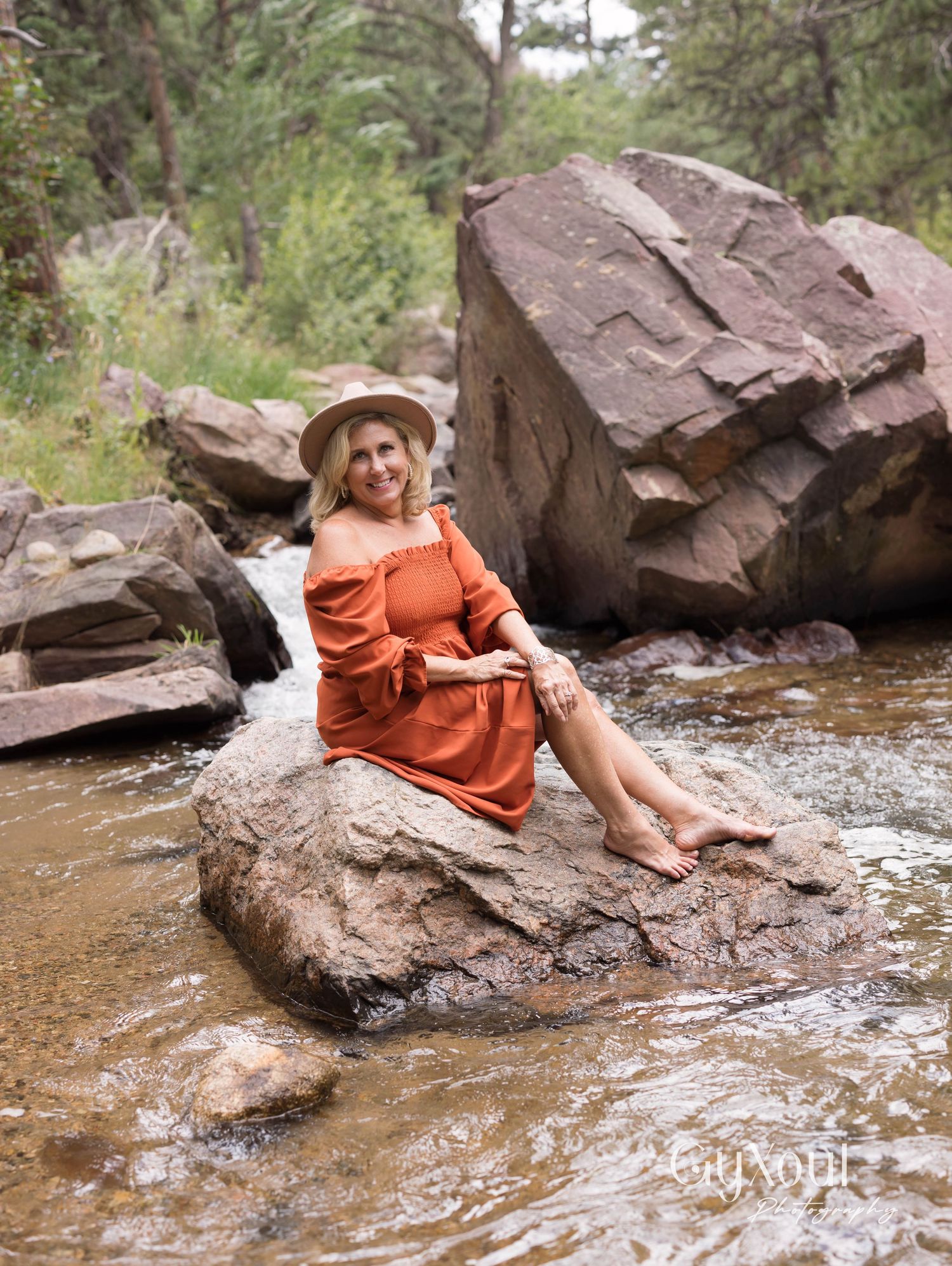 A woman in an orange dress and sun hat sits on a large rock formation by a flowing stream in a wooded area.