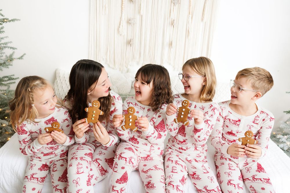 Five children wearing matching white and red pajamas laughing together while holding cookies during the holidays.