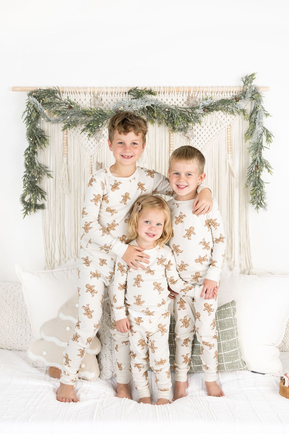 Three children wearing matching gingerbread print pajamas pose in front of a macrame wall hanging with greenery garland.
