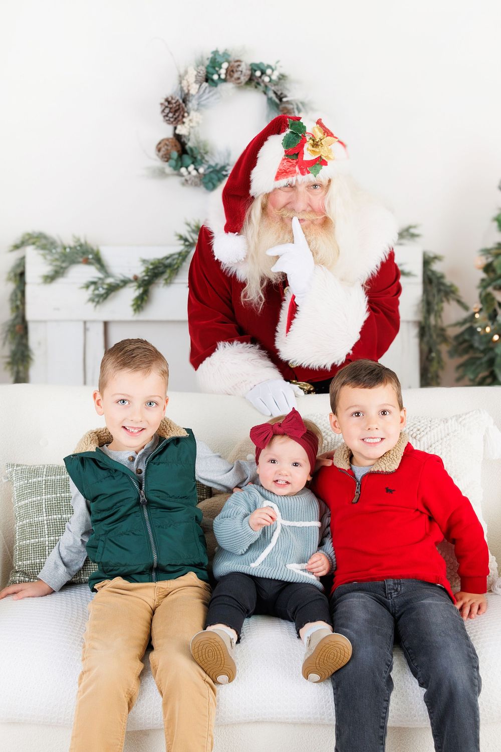 Santa Claus sits on a white couch with three children in front of a winter wreath and greenery backdrop.