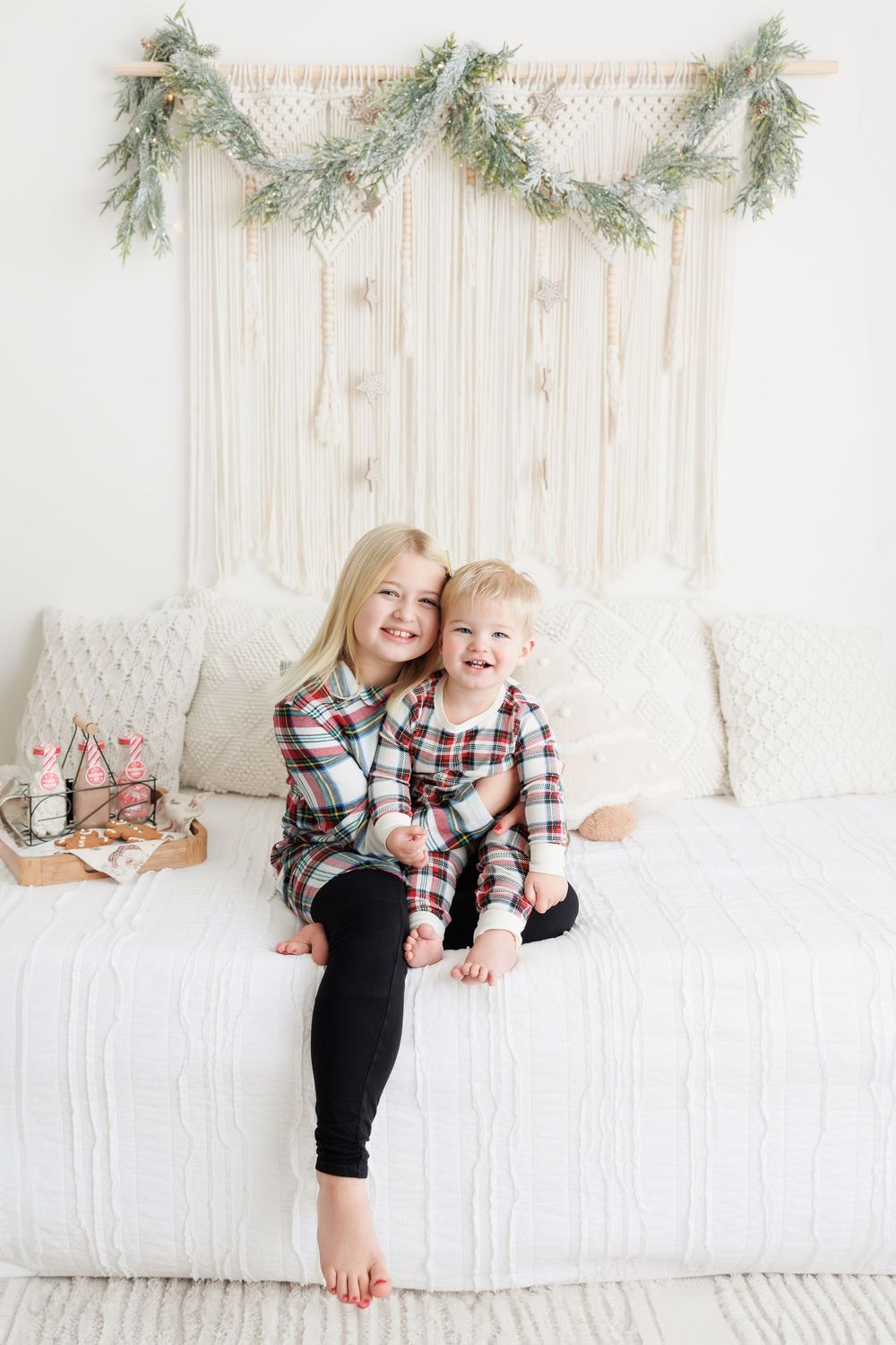 Two children in matching plaid shirts sit together on white couch under evergreen garland wall decor.