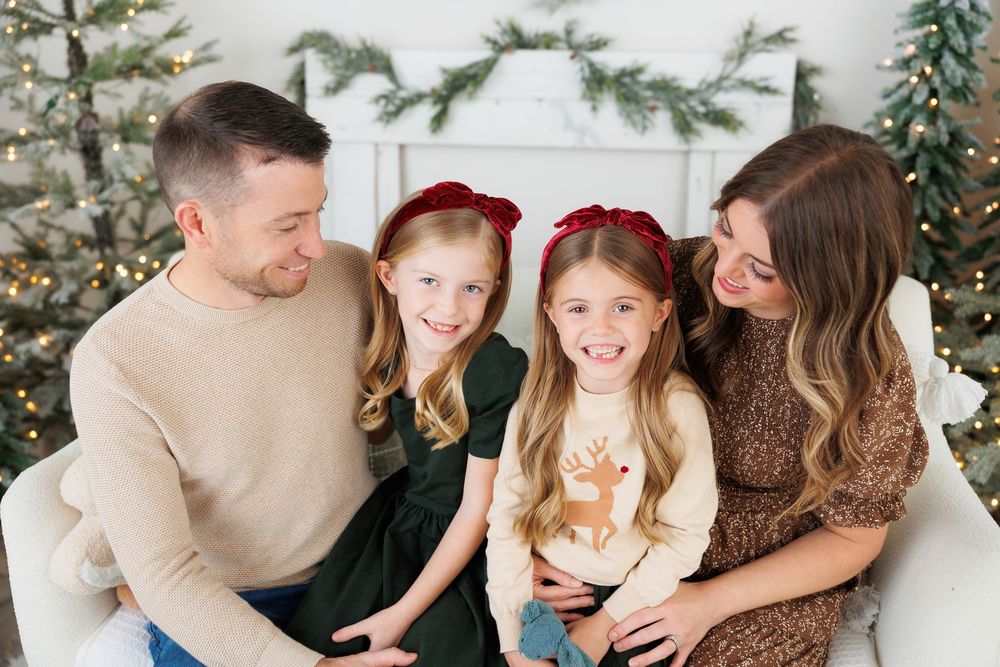 A happy family of four sits together in front of a decorated Christmas tree during a festive holiday photoshoot.