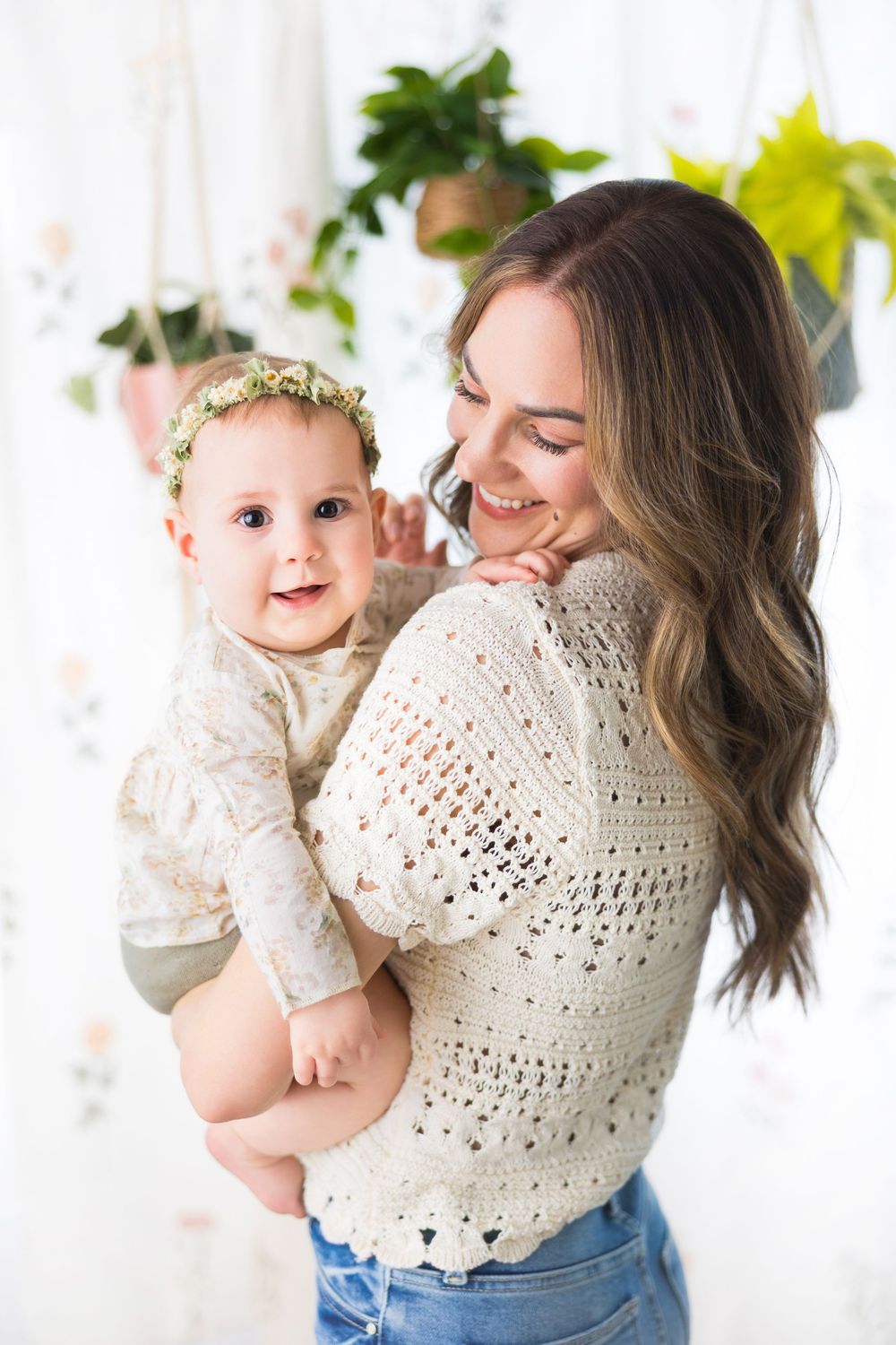 Mother and baby wearing white crochet sweaters embrace in front of green plants and white curtains.