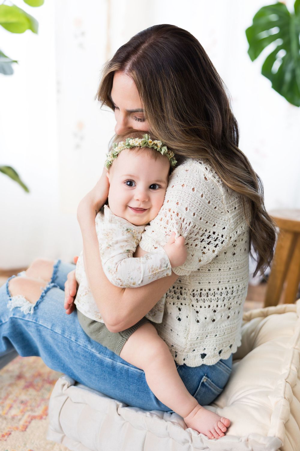 A tender embrace between parent and baby with floral decor and monstera leaves in a bright indoor setting.