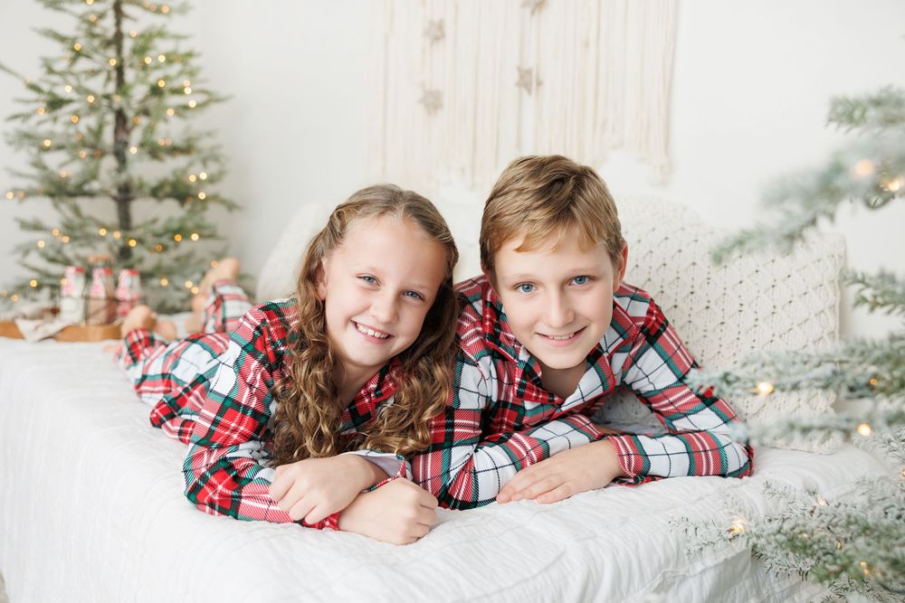 Two children in matching plaid shirts lay together on white bedding with a Christmas tree in the background.