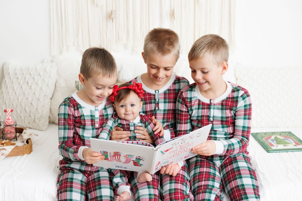 Four children in matching plaid pajamas read a book together on a white couch during holiday season.
