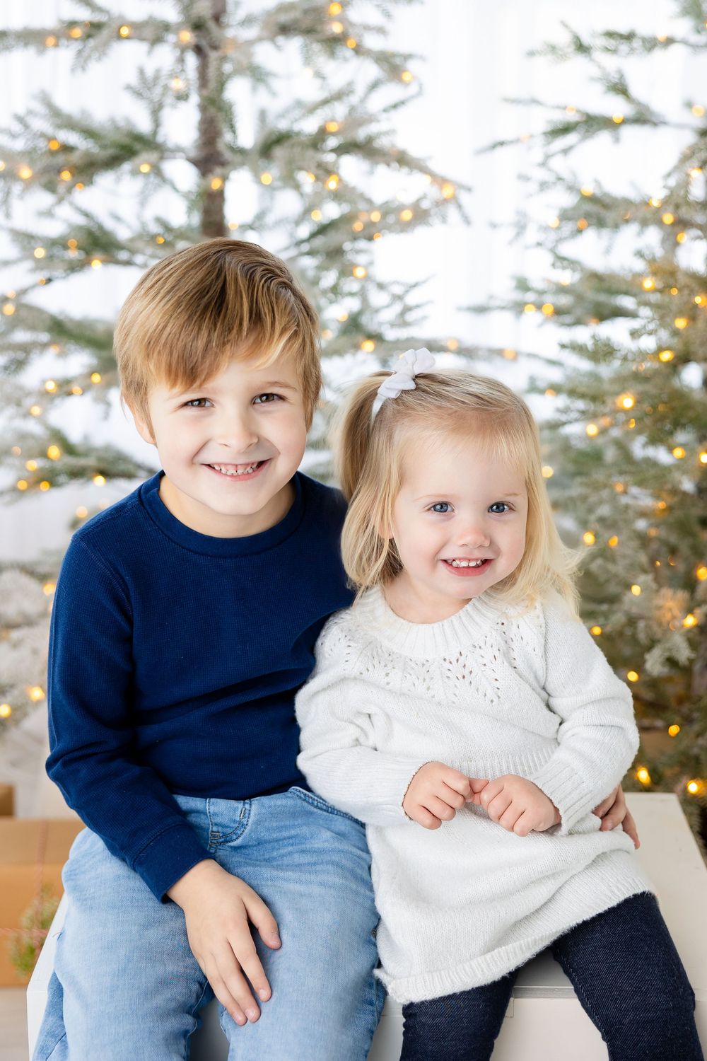 Two young children sit together on a bench smiling in front of a decorated Christmas tree with twinkling lights.