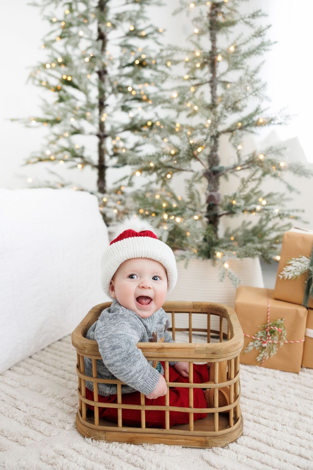 Happy baby wearing a winter hat sits in wooden basket with Christmas tree and wrapped presents in background.