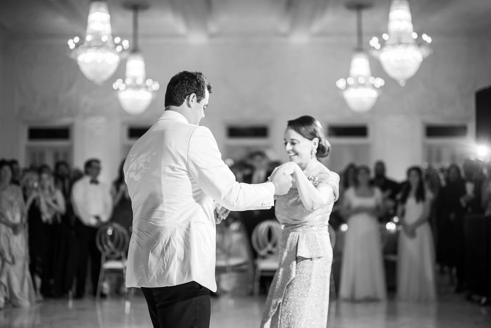 A couple dances gracefully together at a formal event in a ballroom with crystal chandeliers overhead in black and white.