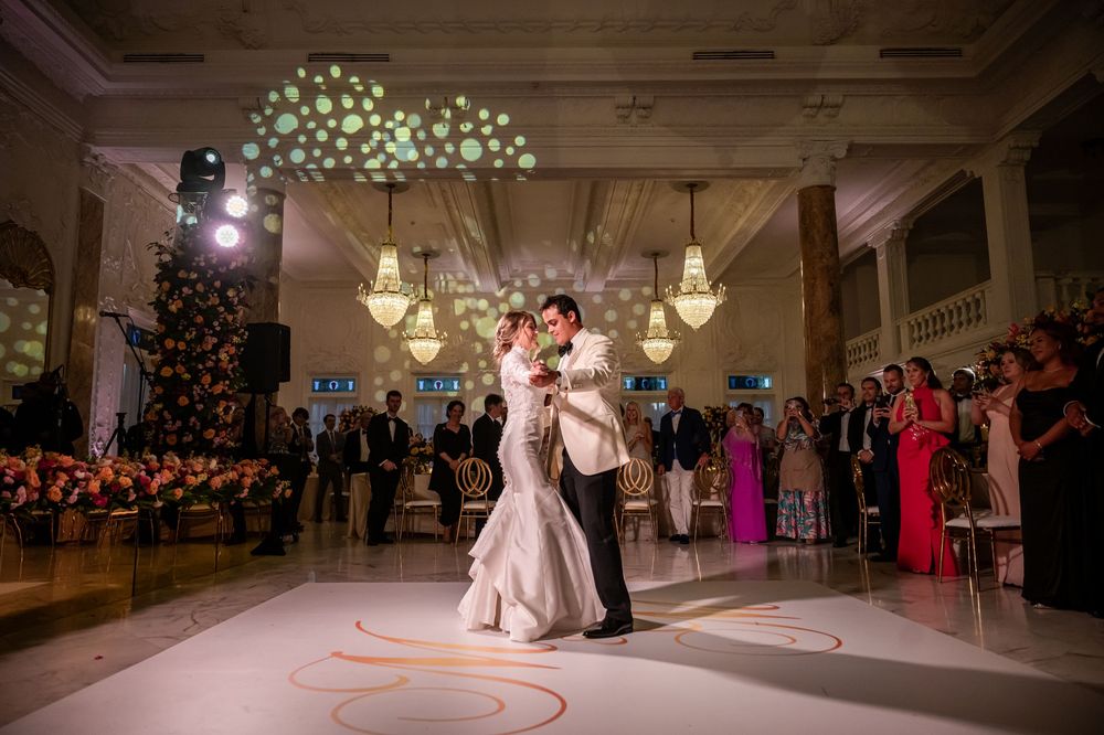 Newlyweds share their first dance under elegant chandeliers with wedding guests watching from the ballroom perimeter.