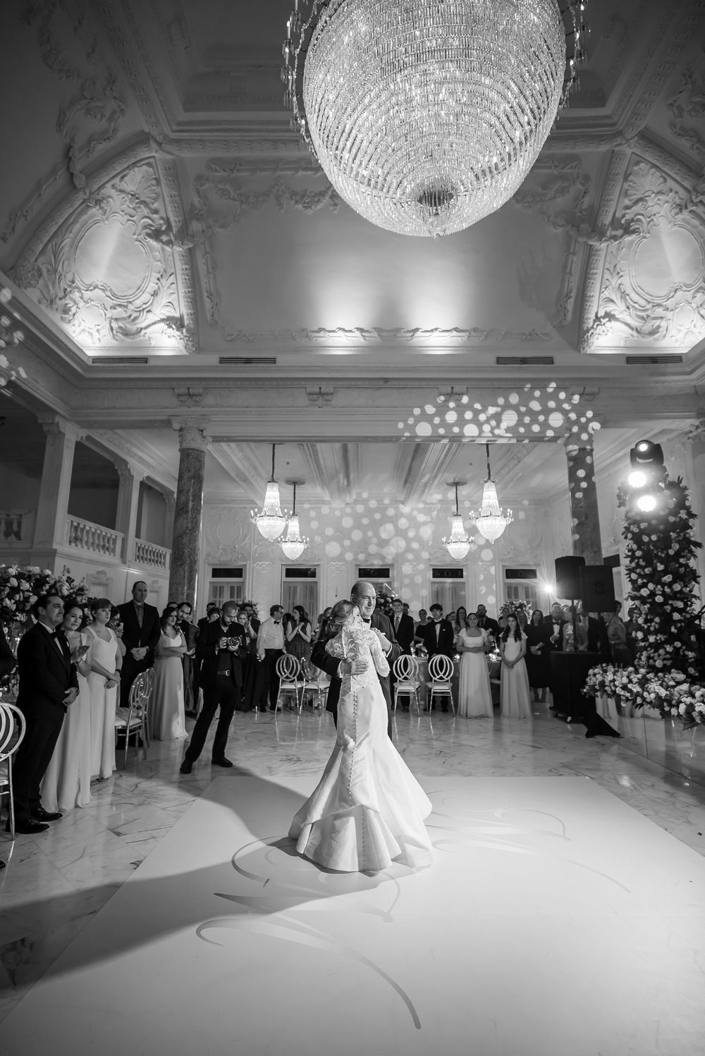 A stunning ballroom wedding reception with a grand crystal chandelier and elegant white dance floor under ornate ceiling details.