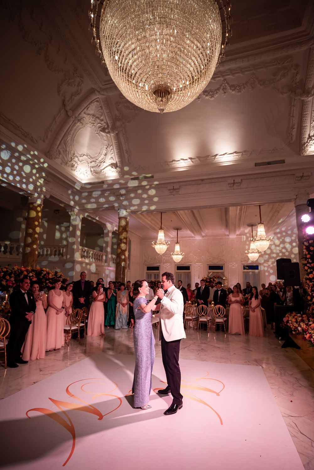 Couple dancing under elegant crystal chandelier in grand ballroom with pink-dressed guests and ornate white decor lighting.