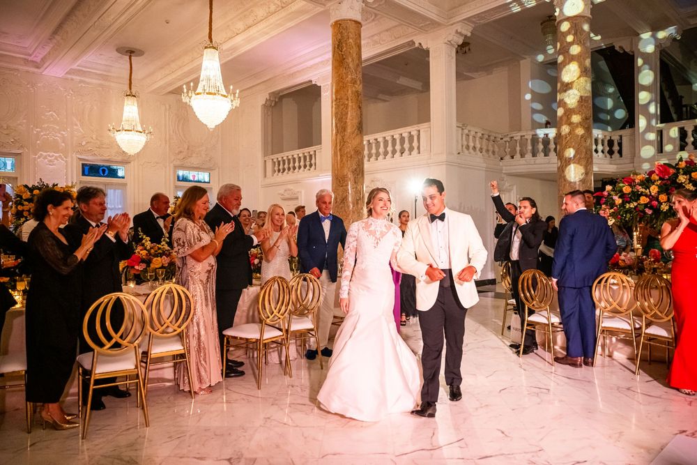 Elegantly dressed couple walks down aisle in grand ballroom with crystal chandeliers and ornate gold chairs.