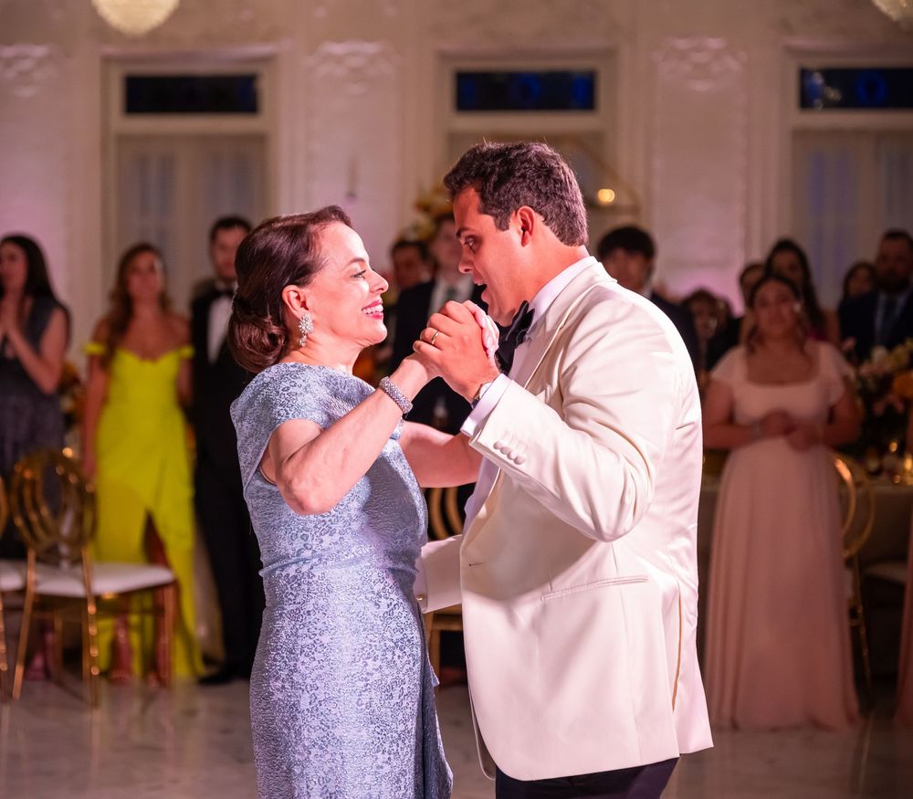 Two people share an emotional dance moment at an elegant reception in a white ballroom with guests in formal attire watching.