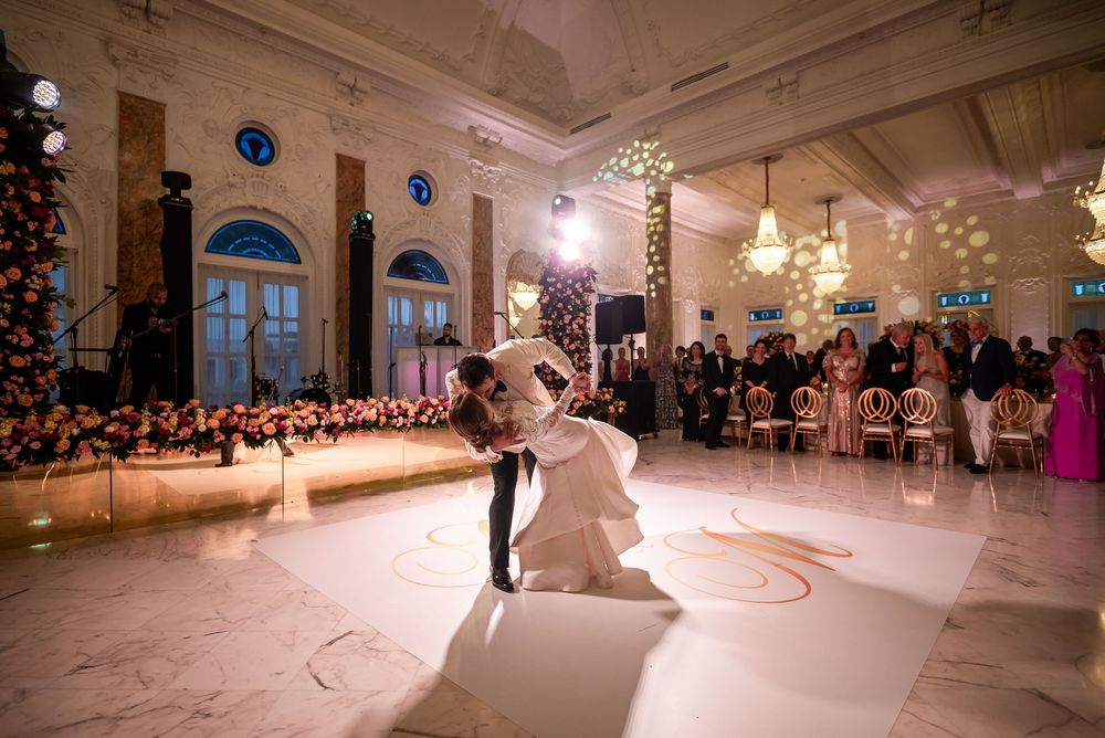 A couple shares their first dance on an elegant white dance floor surrounded by holiday decor in a grand ballroom.