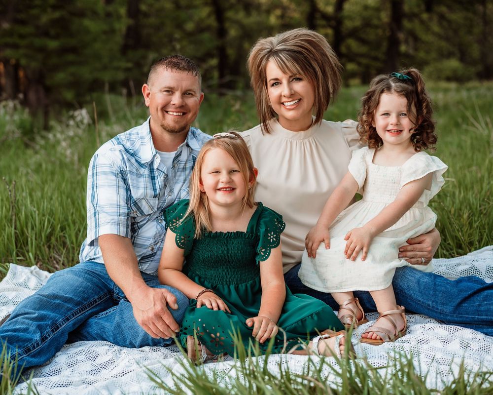 A family of four sits together on a blanket outdoors surrounded by tall grass and pine trees in the background.