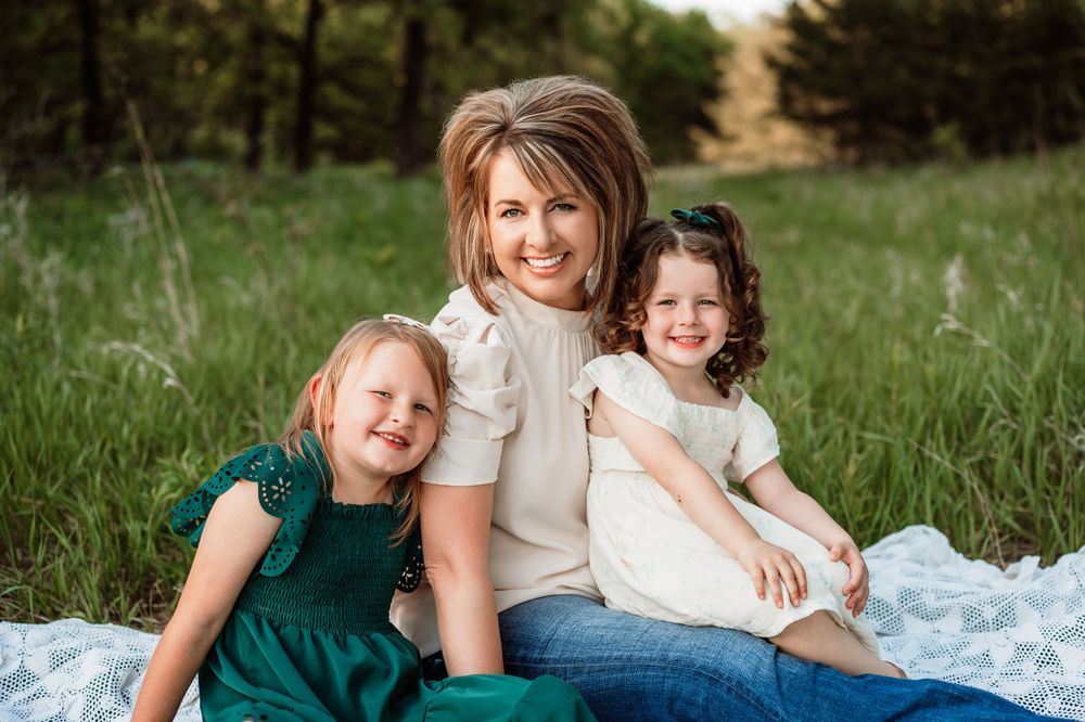 A family sits together on a white blanket in a grassy field during a sunny outdoor photo session.