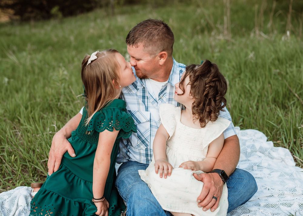 Family sits together on a white blanket in a grassy field wearing coordinated blue, green and white outfits.