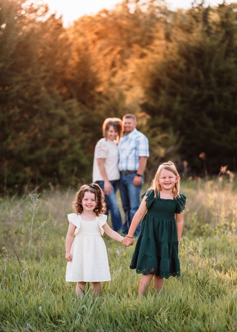 Two children holding hands in a grassy meadow at sunset with family standing in the background.