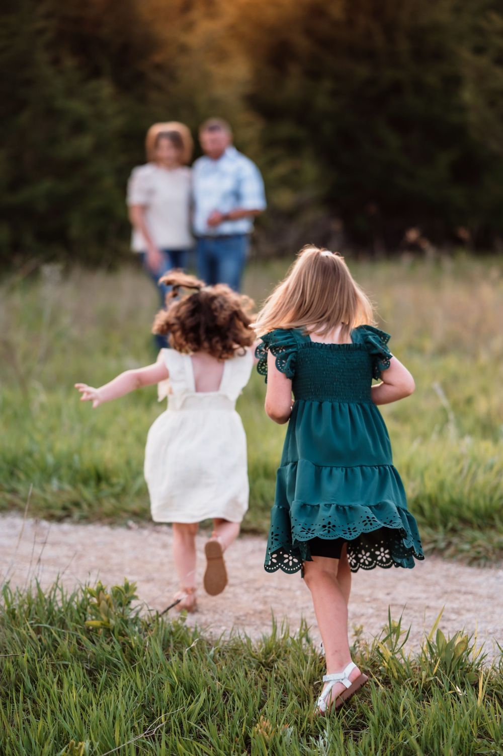Two young children in dresses run along a dirt path through a grassy field on a summer evening.