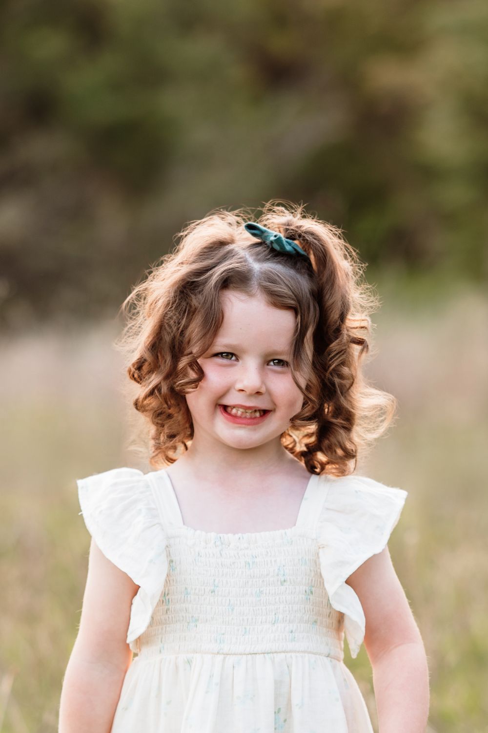 Young child wearing white dress and blue hair bow smiles outdoors with curly hair in natural setting.
