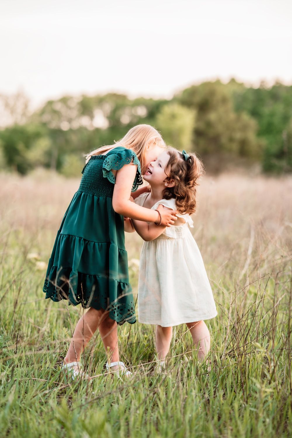 Two children in dresses share a sweet embrace while playing together in a tall grass meadow at sunset.