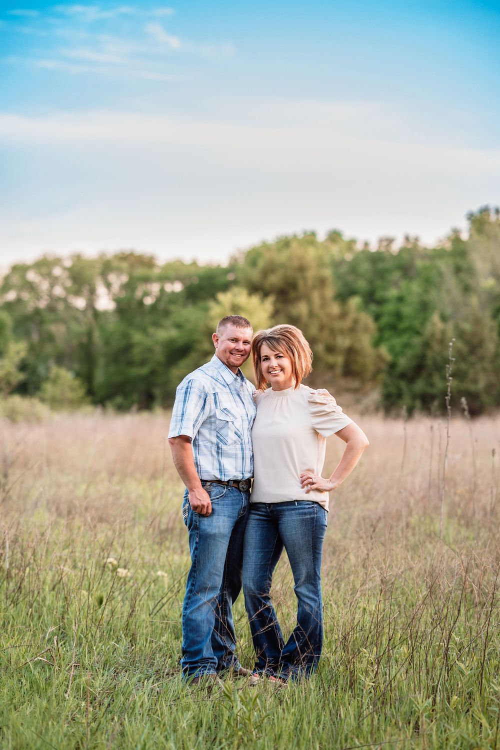 A couple stands together in a sunlit meadow with trees in the background during golden hour.