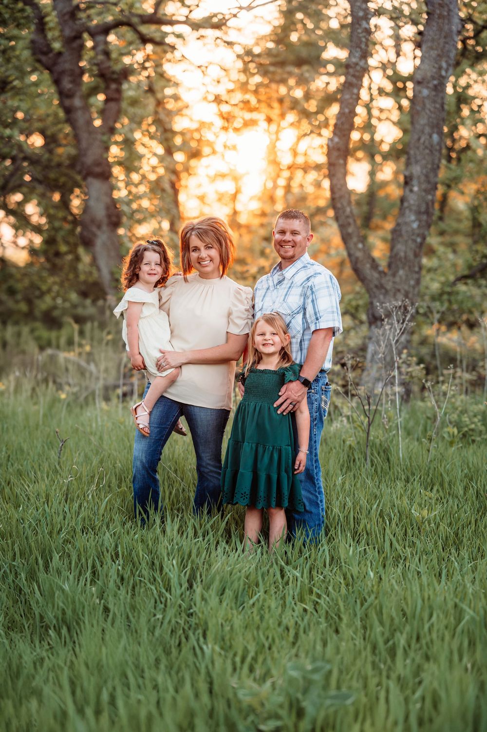 A family stands together in a grassy field at sunset with trees in the background creating a warm golden atmosphere.