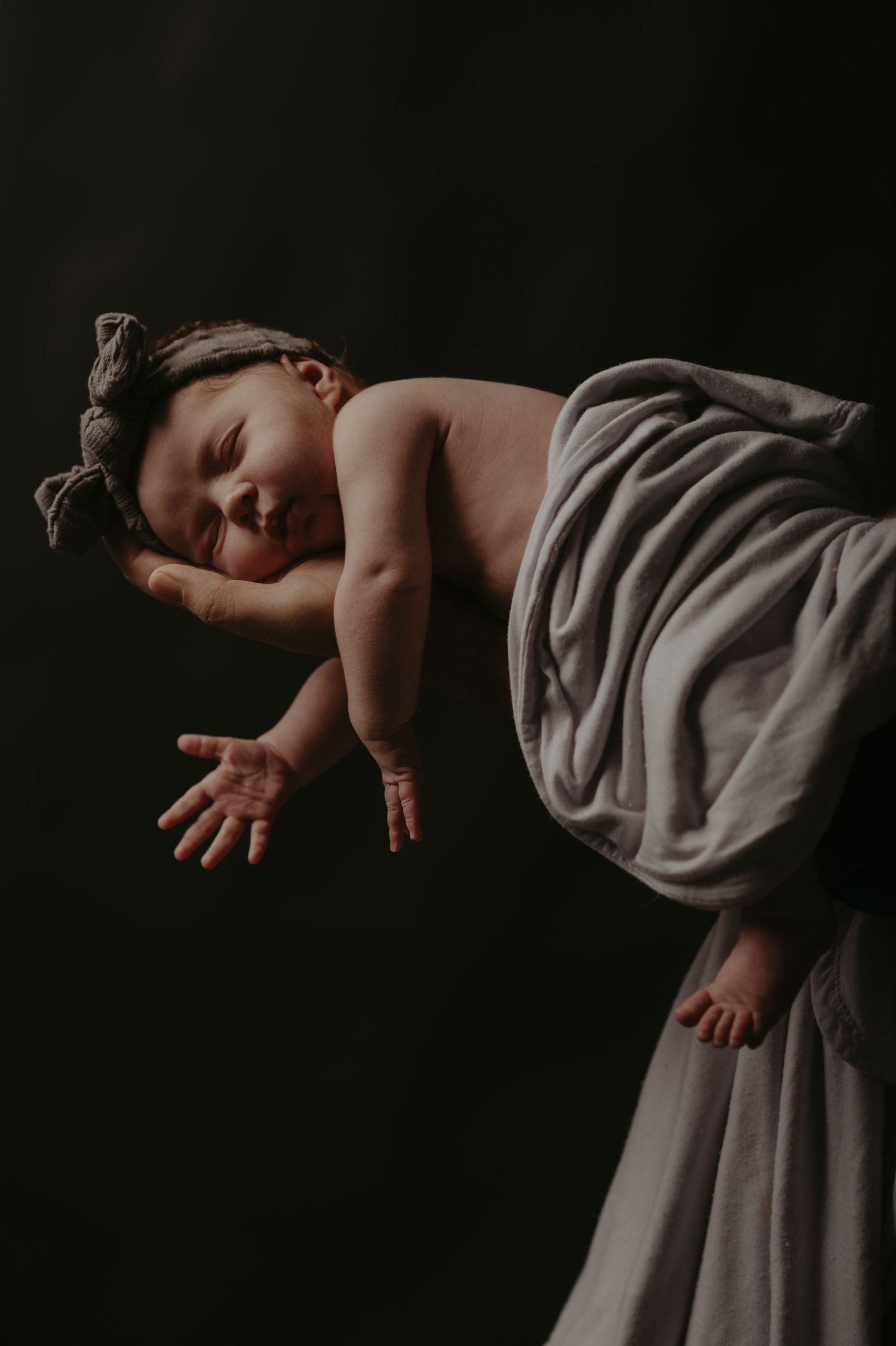 Sleeping baby wrapped in soft gray fabric against dark background in artistic studio portrait.