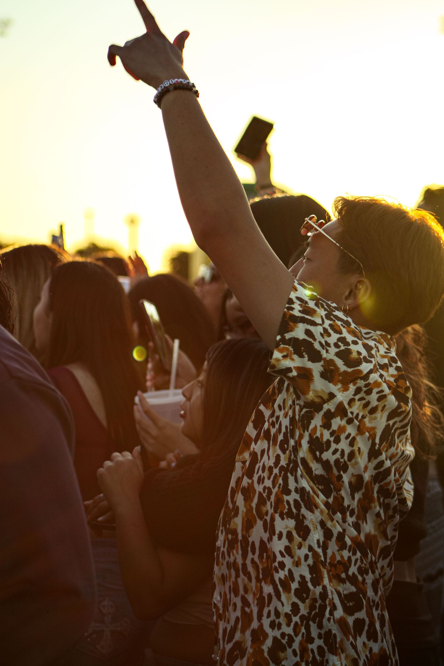 Concert fan in leopard print shirt raising hand at outdoor music festival during sunset.