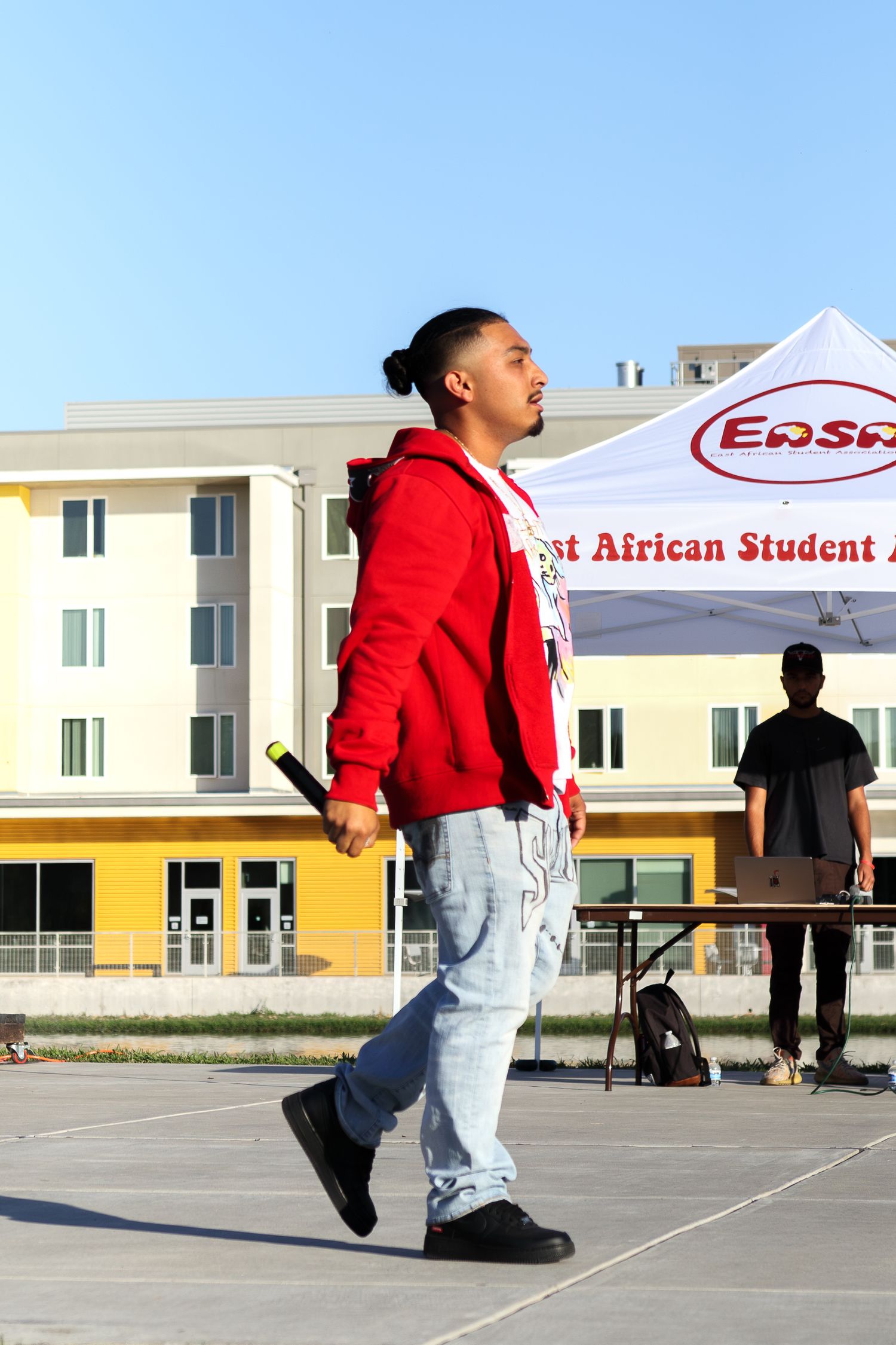 Young man in red hoodie and jeans walking near East African Student Association tent outdoors.