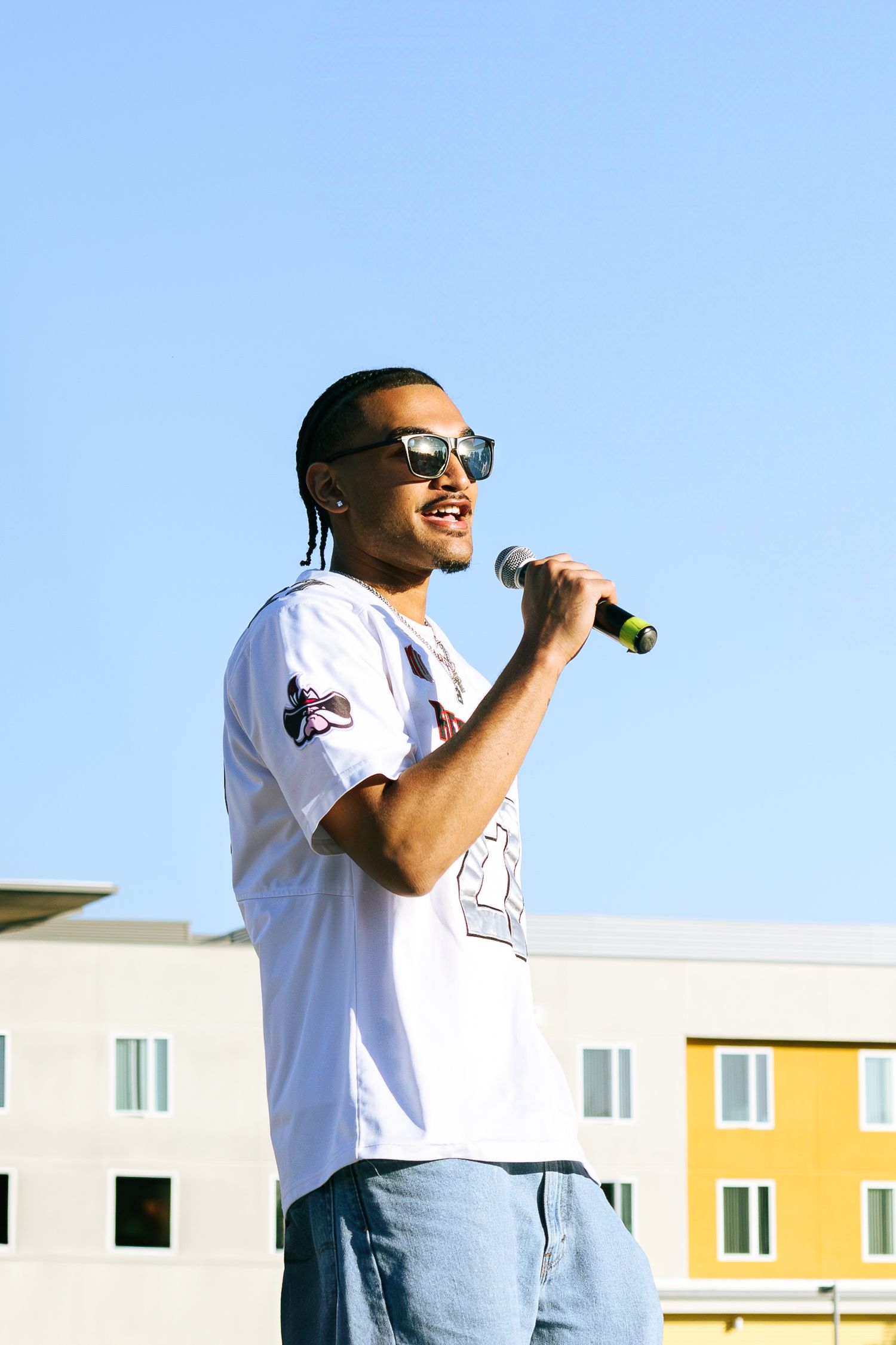 Young man in white t-shirt and sunglasses performing with microphone at outdoor event.