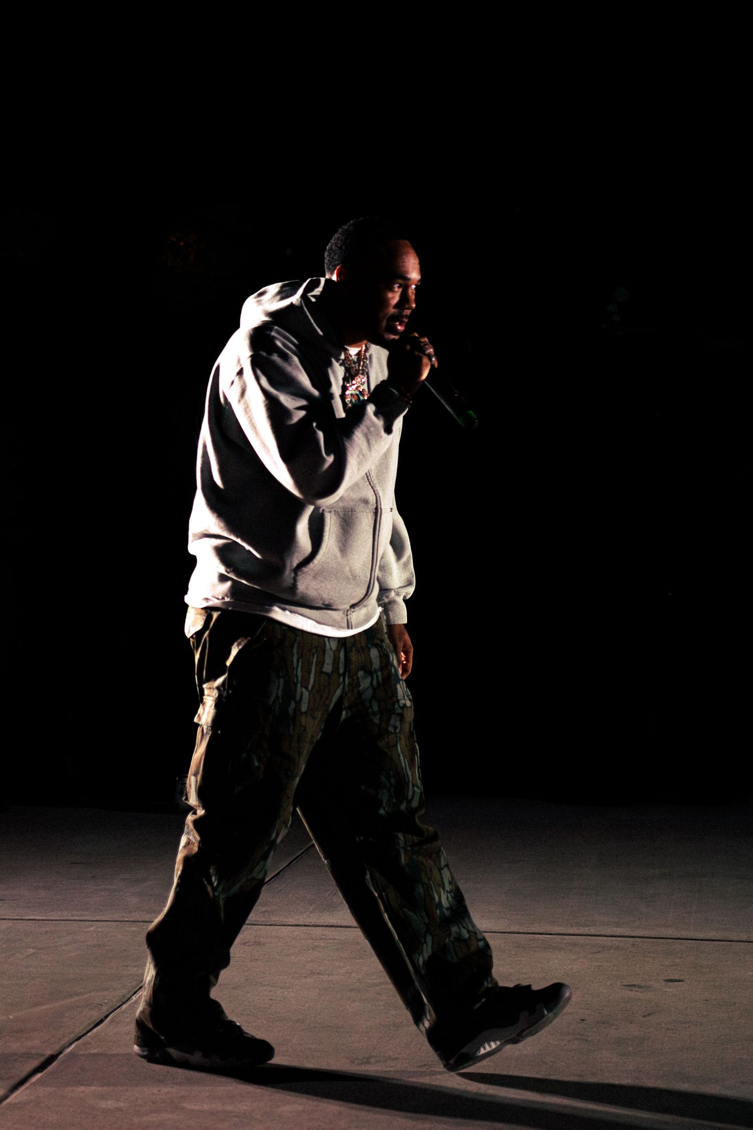 A male rapper performing on stage in a gray hoodie and camouflage cargo pants, holding a microphone in dark spotlight.