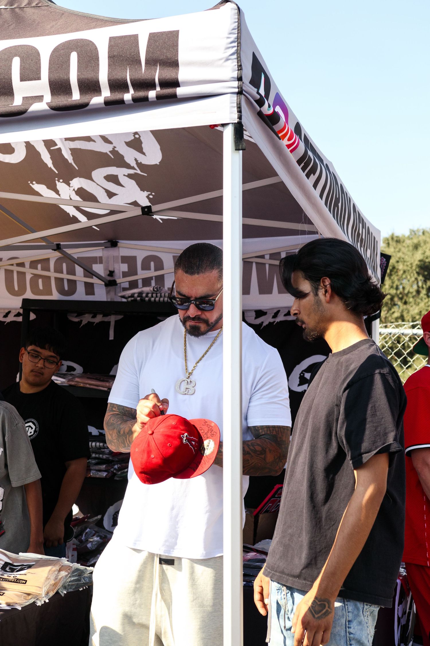 Tattooed man in white shirt signing a red cap for a fan at an outdoor vendor booth.