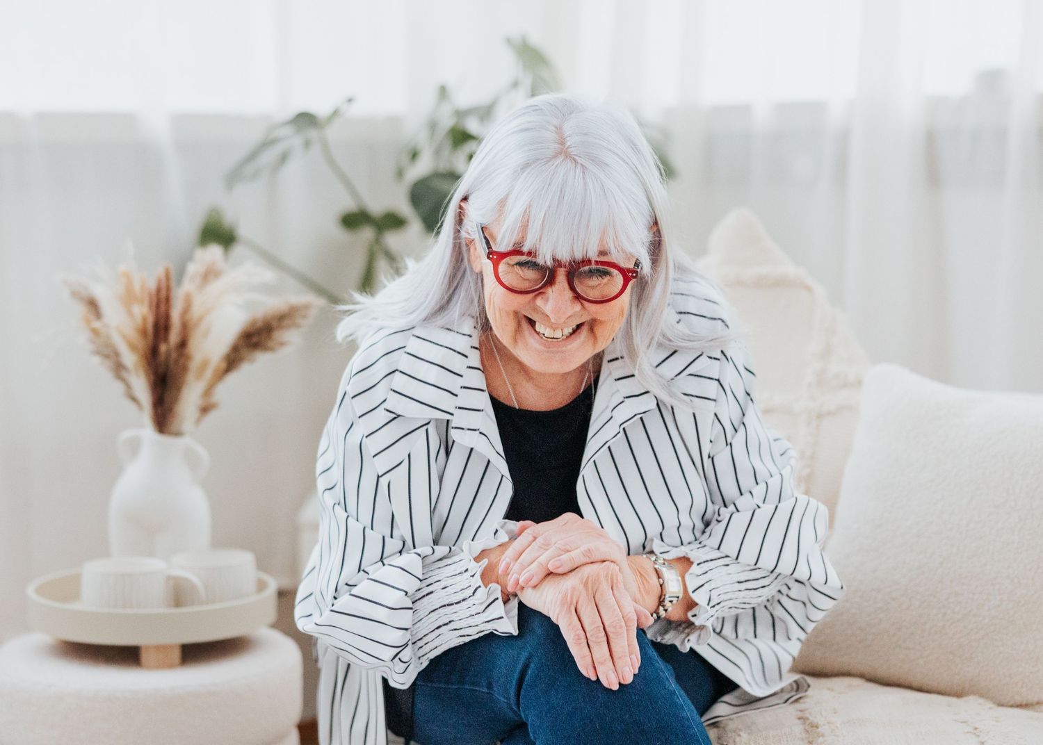 Someone wearing a striped blazer and glasses sits on a white couch while laughing