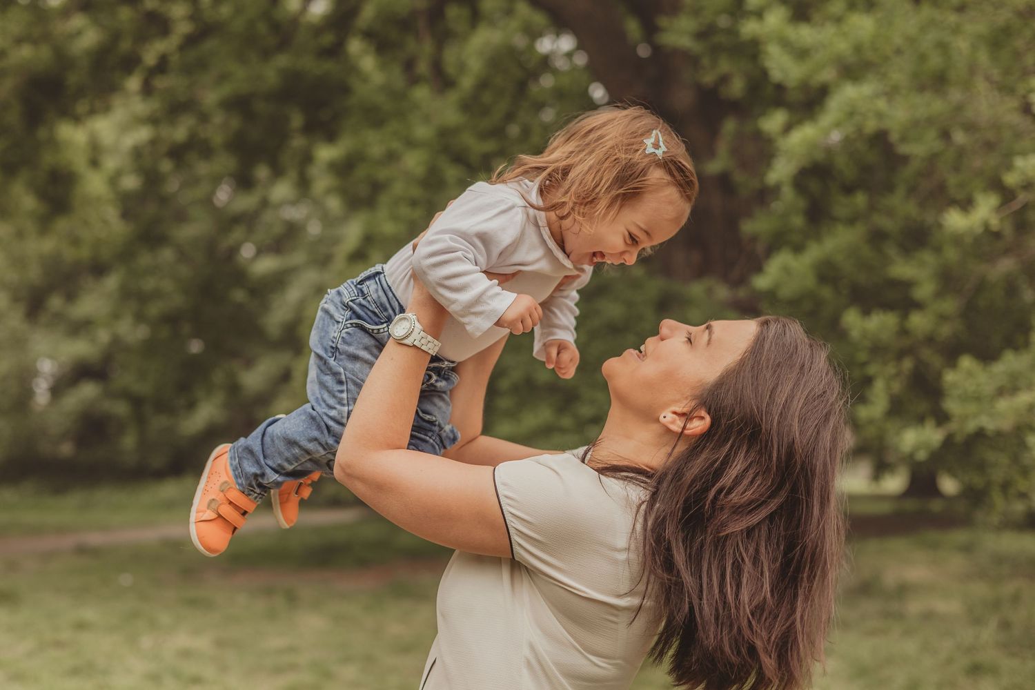 A fun image of a mother playfully lifting her toddler daughter into the air as they both smile and laugh, captured in the green outdoors