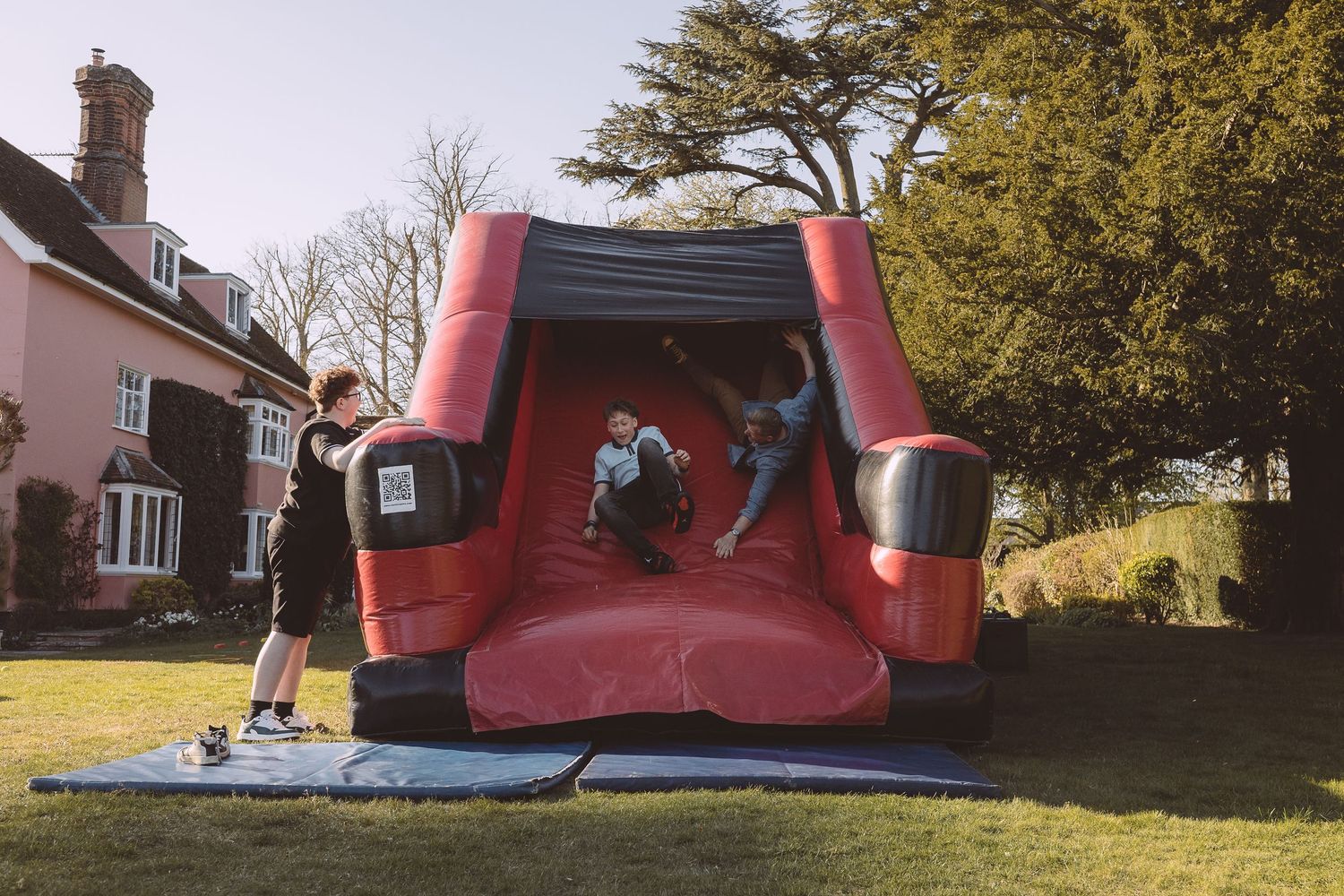Large gathering in a sunny garden with a pink Victorian house and red inflatable bouncy castle slide in the background.