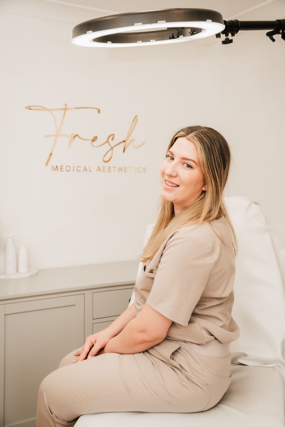 Medical aesthetician in beige scrubs sits on treatment chair under ring light at Fresh Medical Aesthetics clinic.