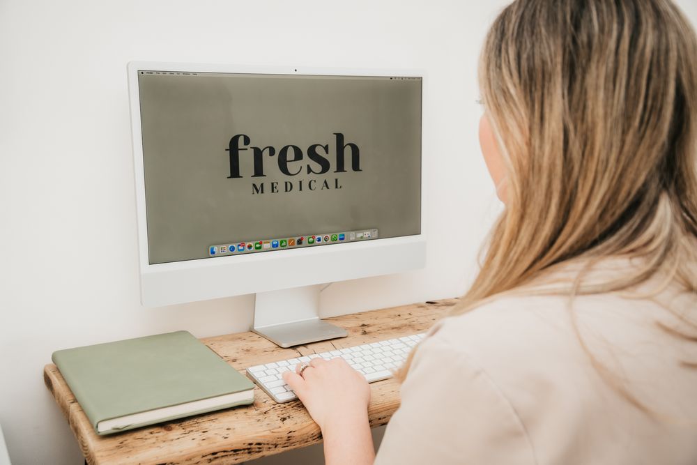 Someone in a beige sweater working at a wooden desk with a computer monitor displaying text that says 'fresh'.