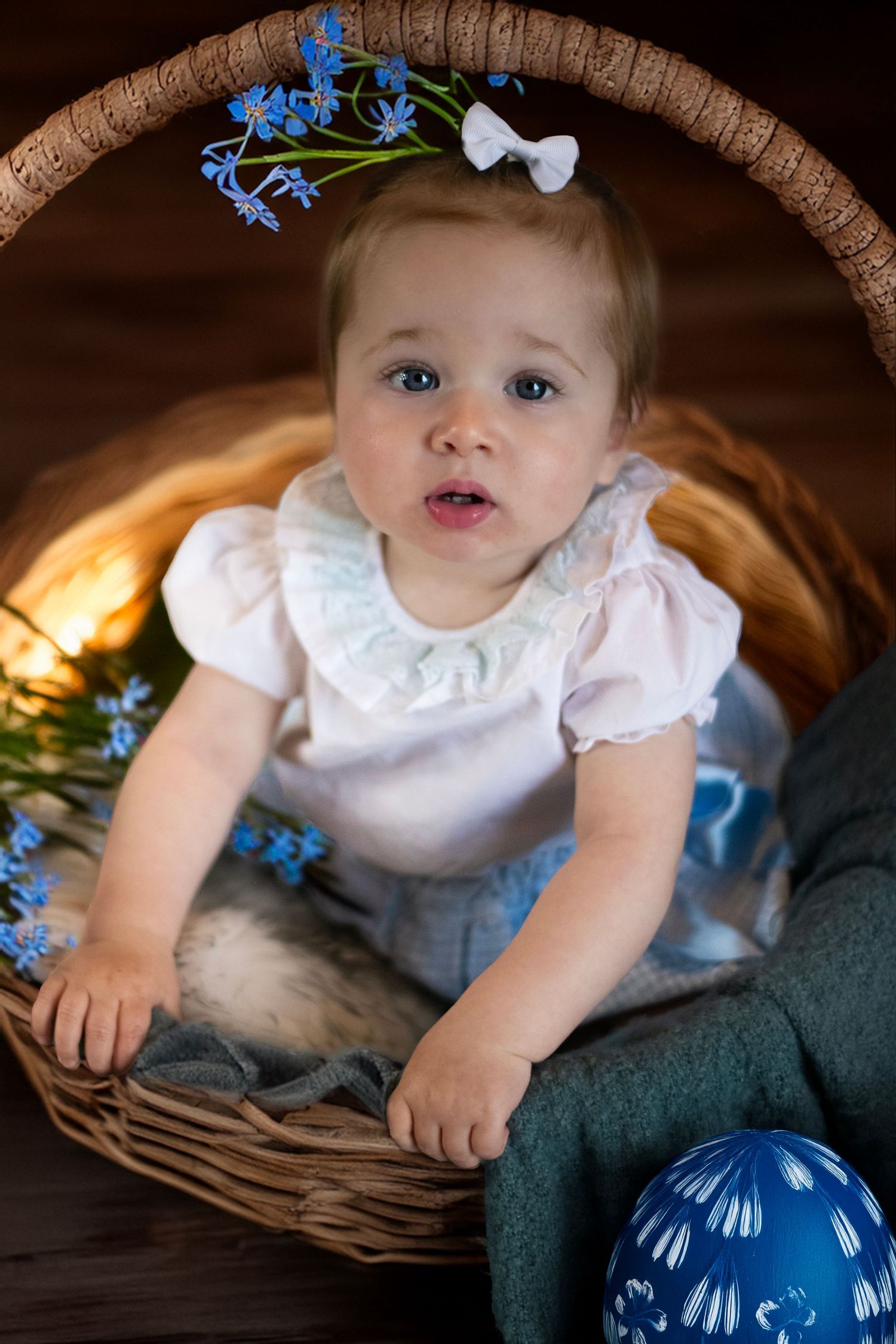 Baby in white dress sits in wicker basket with blue flowers and decor against dark background.