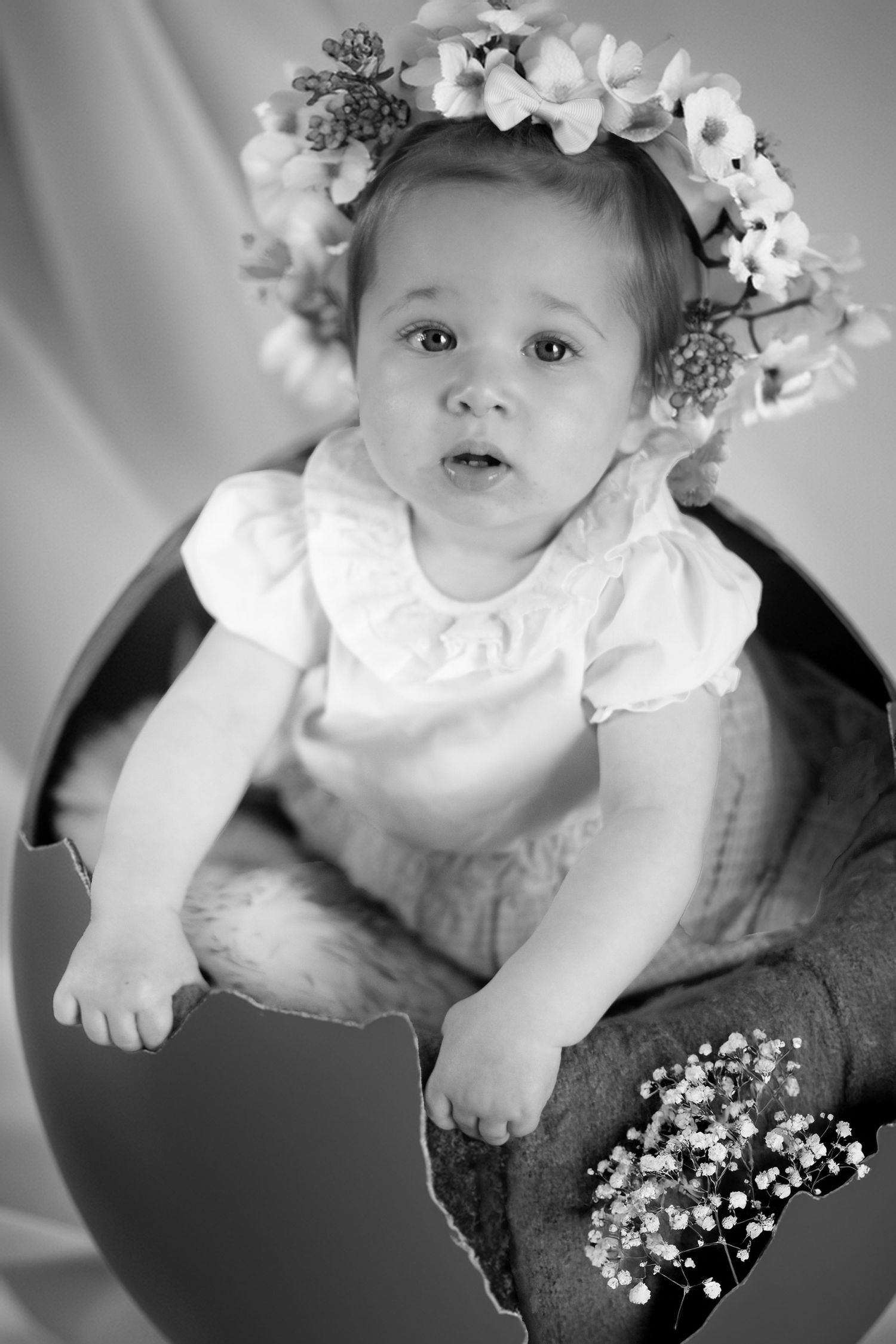 Black and white portrait of a baby wearing a white dress and floral crown sitting on a dark cushion.