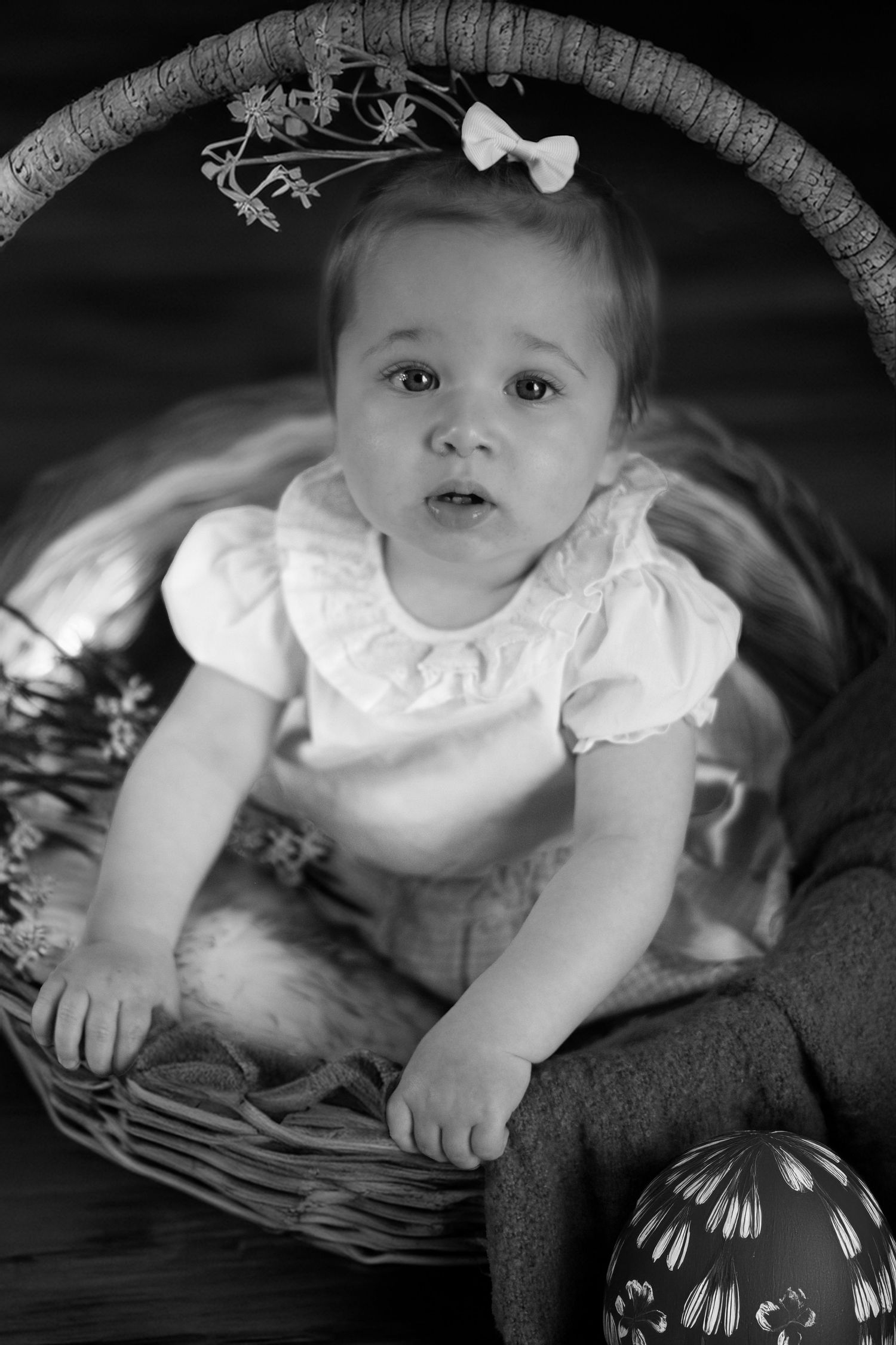 Black and white portrait of a baby in a white dress sitting in a woven basket with a small bow accessory.