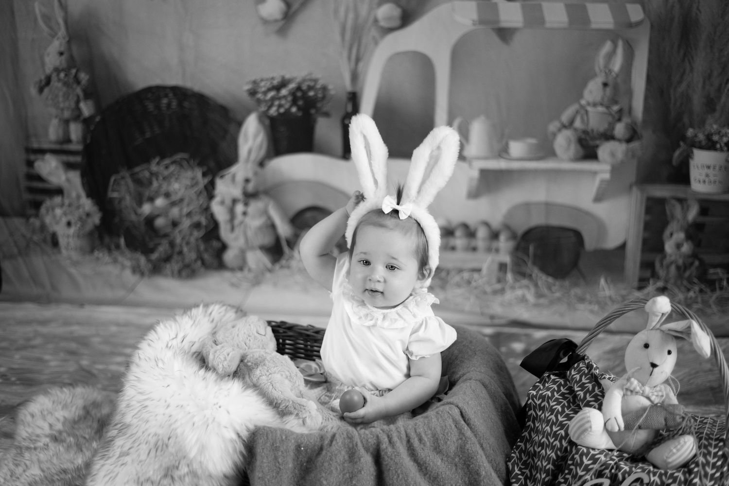 A vintage black and white photograph of a baby wearing bunny ears while sitting on a plush cushion with toys nearby.