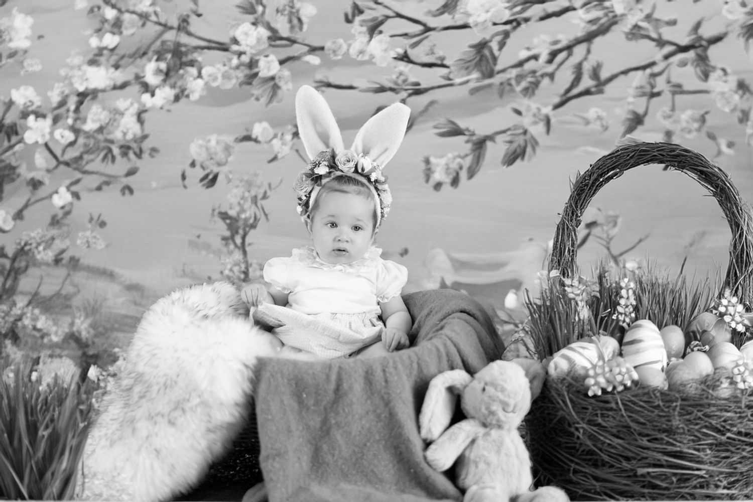 Black and white Easter photo with bunny ears, basket of flowers, and plush rabbit on vintage floral backdrop.