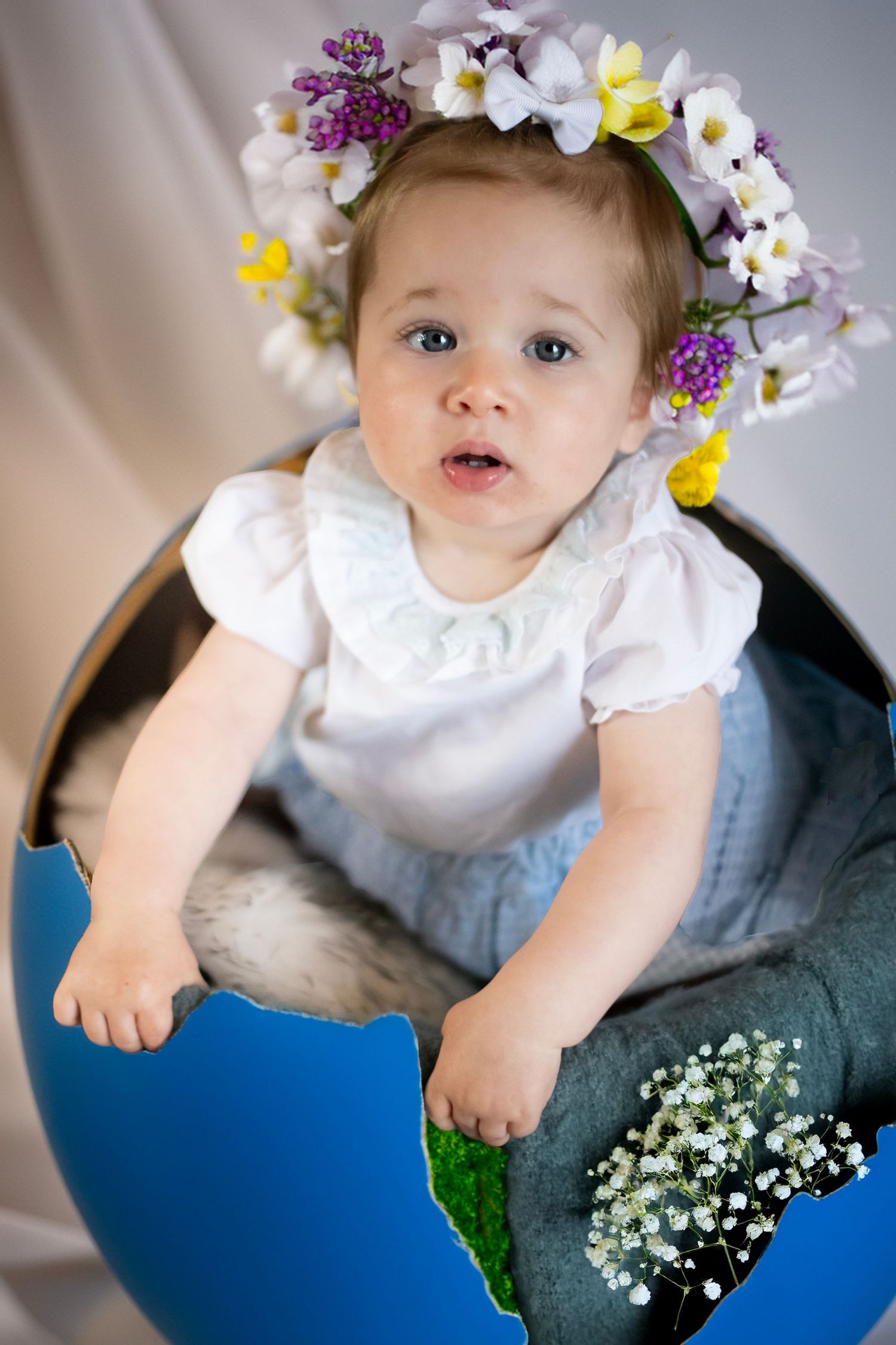 Baby wearing floral crown and white dress poses in blue setting with delicate spring flowers.