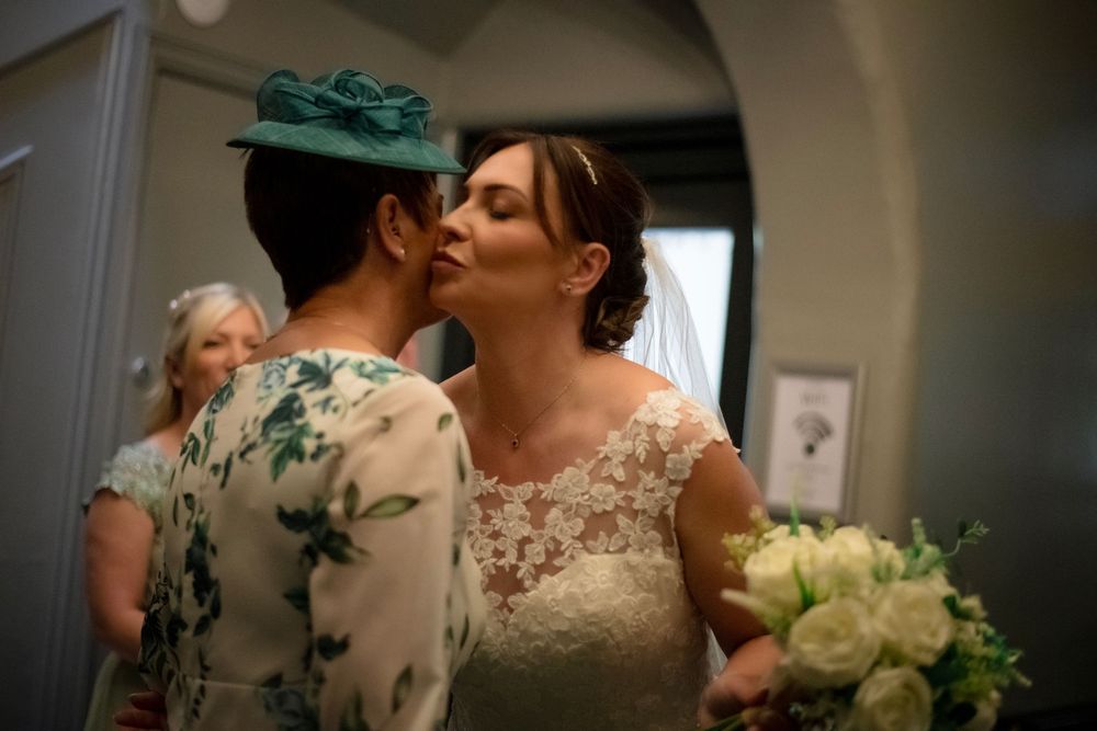 A bride in a lace wedding dress embraces a guest wearing a green floral dress at an indoor venue.
