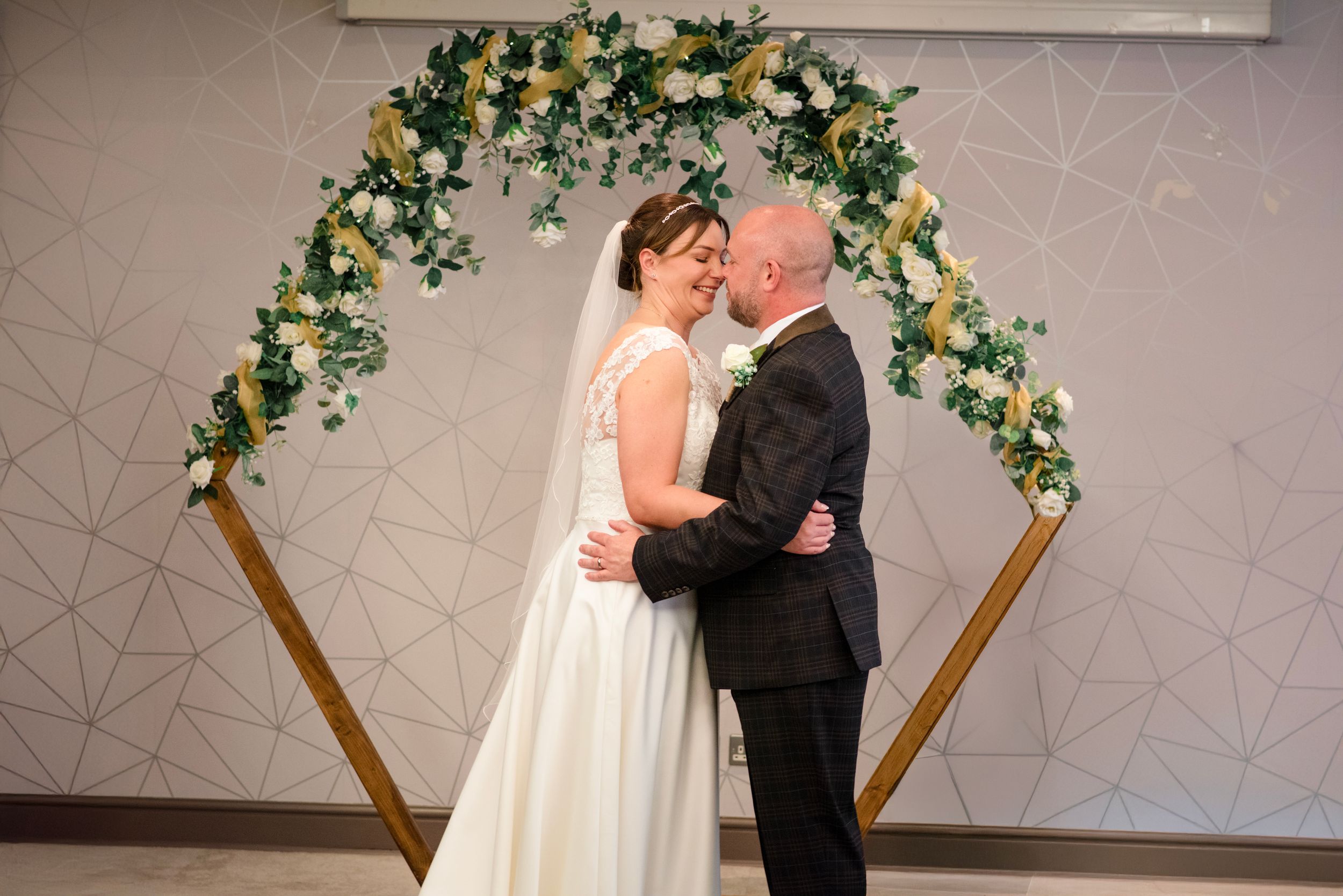 A couple shares their first kiss under an elegant floral arch with white and yellow blooms at their wedding ceremony.
