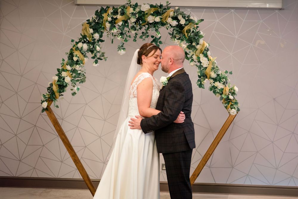 A couple shares their first kiss under an elegant floral arch with white and yellow blooms at their wedding ceremony.