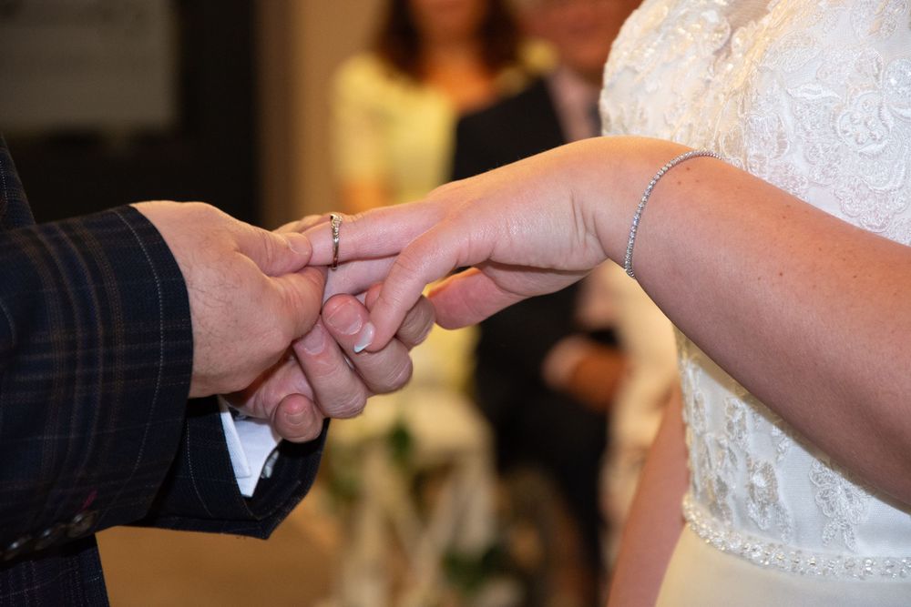 Close-up photo of hands exchanging wedding rings during a marriage ceremony with white lace dress visible.